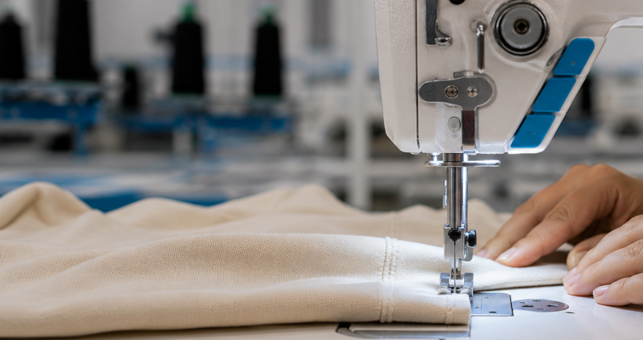 A close-up of a beige fabric being sewn by an industrial sewing machine, with a person's hands guiding the material. The background shows a softly blurred textile workshop with other sewing machines and spools of thread.